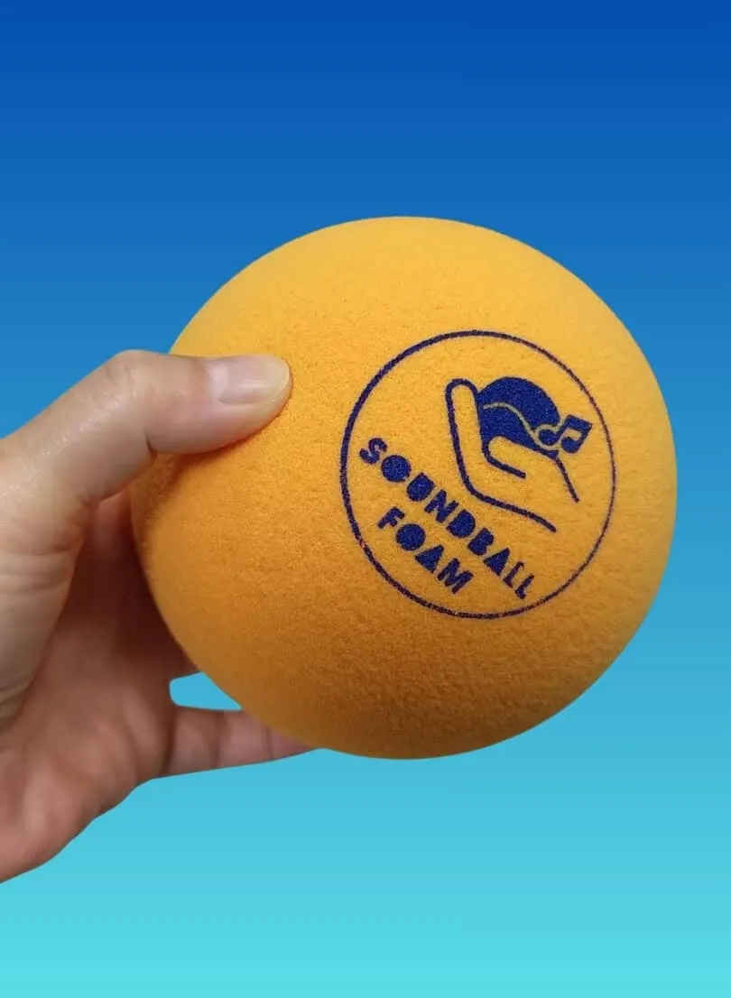 A hand holding a bright orange foam sound ball marked “Soundball Foam”.