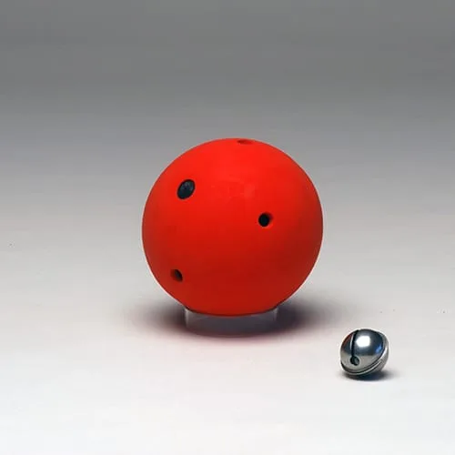 A solid red rubber ball with four dark sound holes drilled through it, with a chrome jingle bell beside it.
