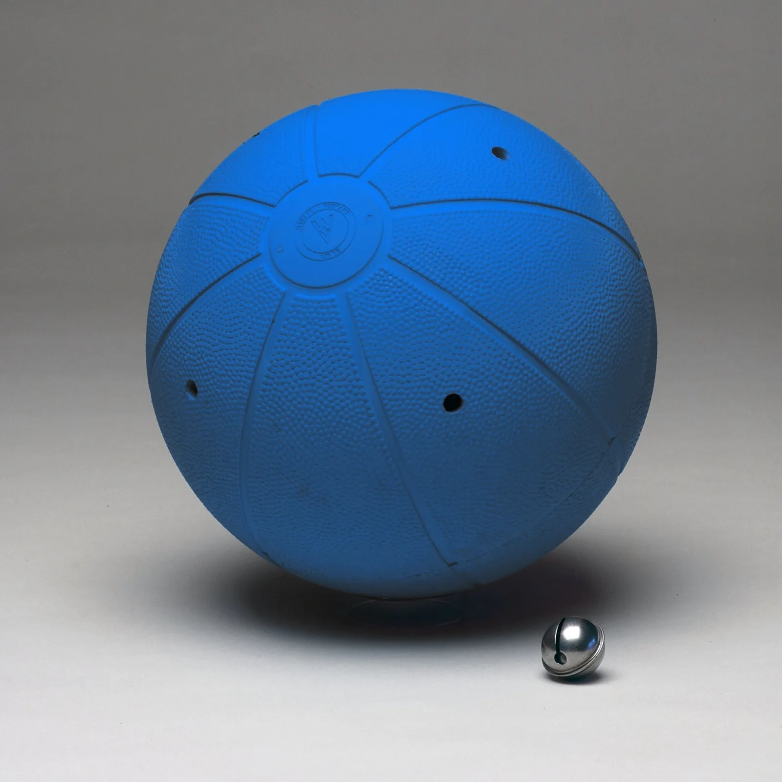 A blue rubber goalball with a pebbled surface, moulded seams and small sound holes, with a chrome jingle bell beside it.